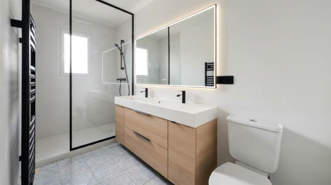 Salle de bain moderne &eacute;pur&eacute;e avec douche &agrave; l'italienne, paroi en verre &agrave; cadre noir, meuble vasque double suspendu en bois clair avec plan vasque blanc int&eacute;gr&eacute;, robinetterie noire mate, grand miroir LED r&eacute;tro&eacute;clair&eacute;, WC &agrave; r&eacute;servoir apparent, s&egrave;che-serviettes &eacute;lectrique noir, carrelage gr&egrave;s c&eacute;rame gris clair, murs blancs