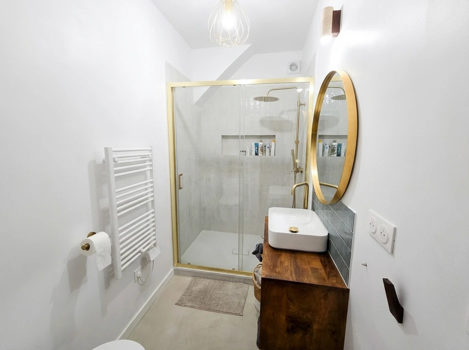 Salle de bain moderne épurée, murs blancs, douche à l'italienne avec paroi coulissante en verre et profilés dorés, receveur blanc, colonne de douche pluie laiton brossé