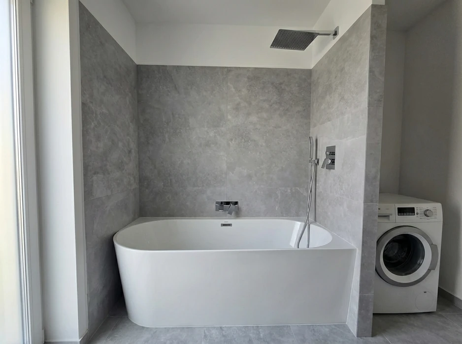 Salle de bain moderne avec baignoire îlot rectangulaire blanche en solid surface, encastrée contre un mur en grands carreaux effet marbre gris