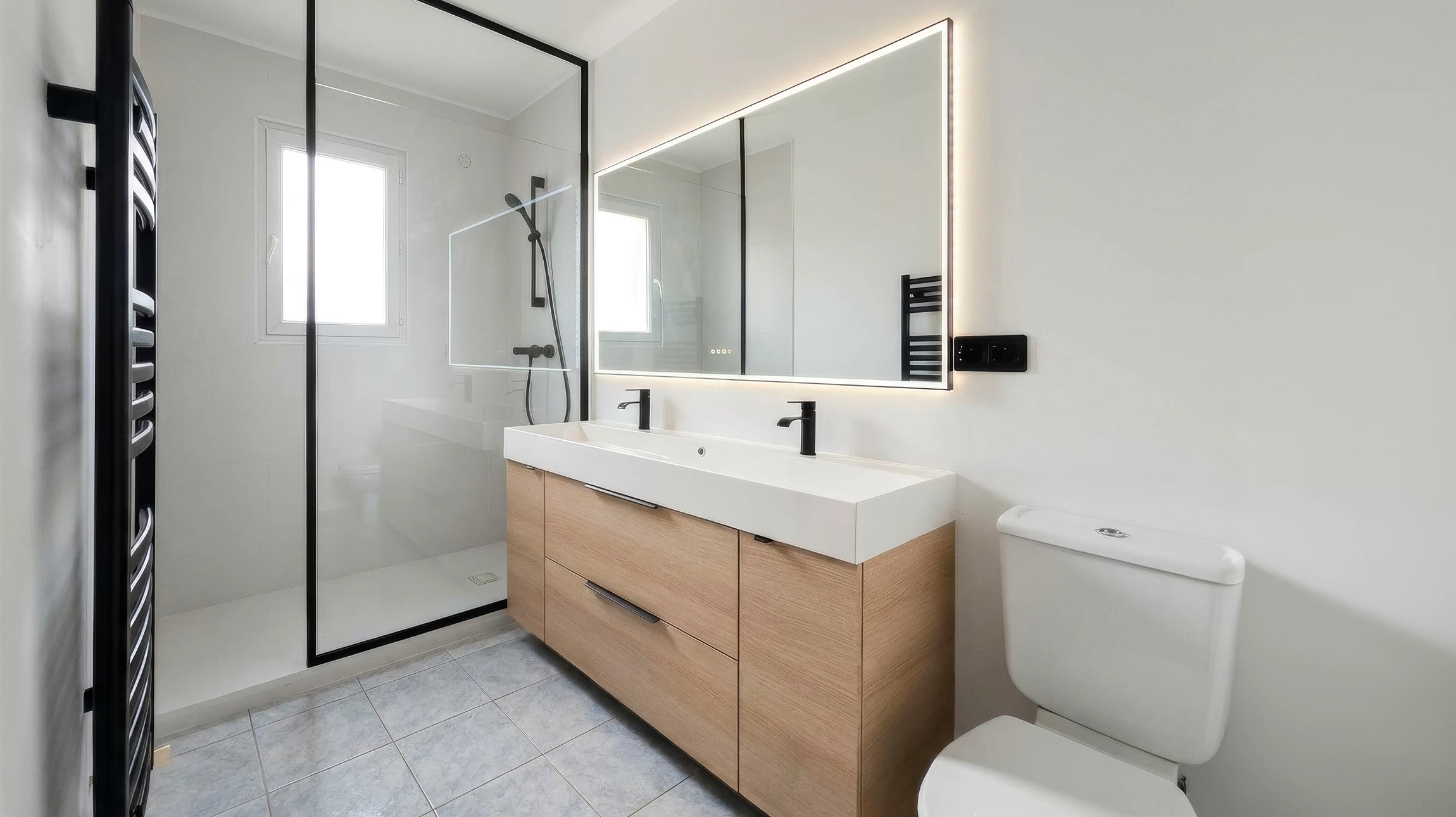 Salle de bain moderne épurée avec douche à l'italienne, paroi en verre à cadre noir, meuble vasque double suspendu en bois clair avec plan vasque blanc intégré, robinetterie noire mate, grand miroir LED rétroéclairé, WC à réservoir apparent, sèche-serviettes électrique noir, carrelage grès cérame gris clair, murs blancs